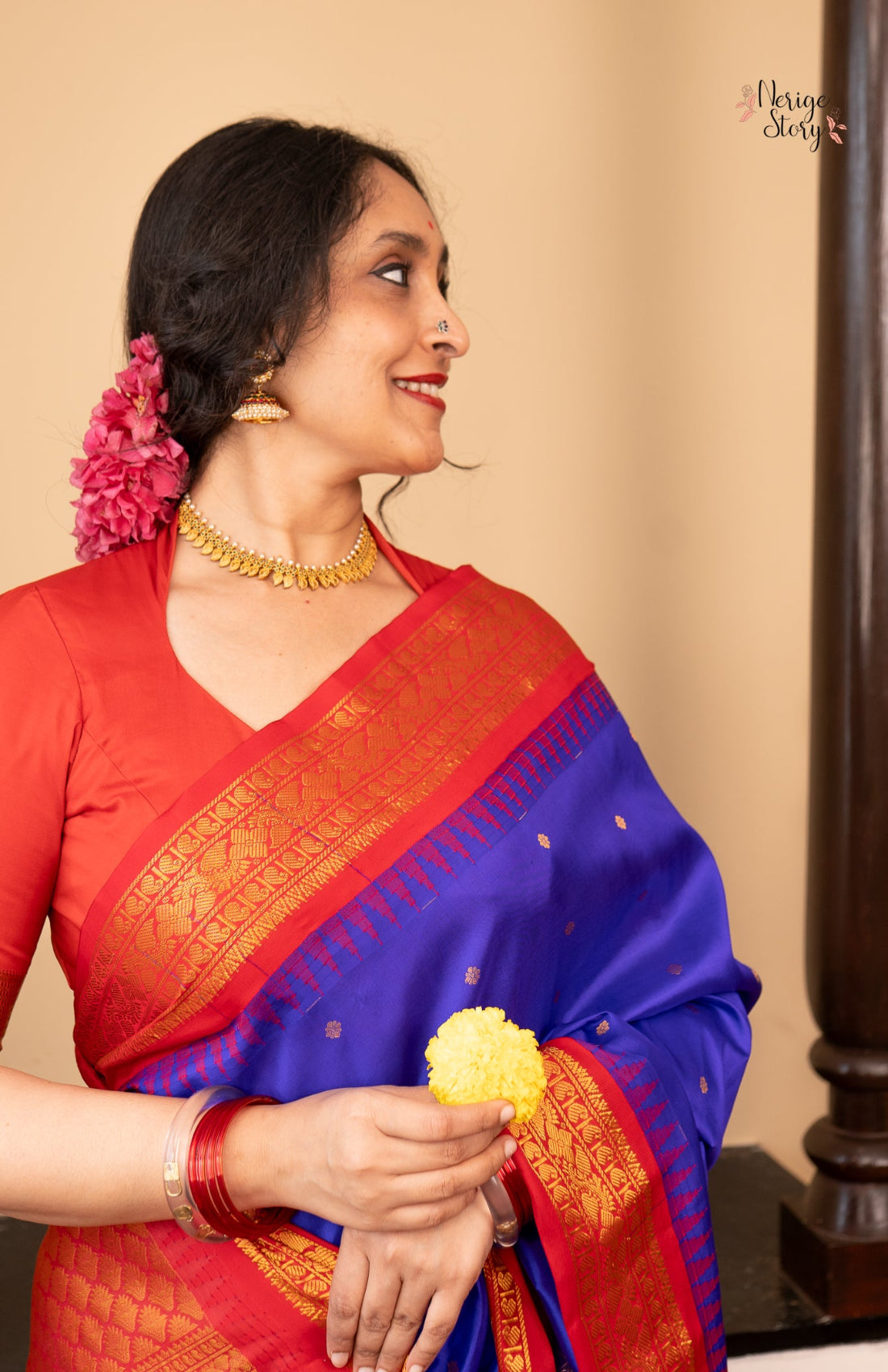 Woman wearing a red and blue saree with a floral decoration, holding a yellow flower against a beige background.