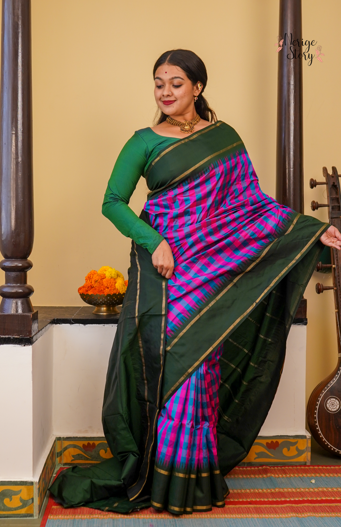 Woman in a green and purple saree standing in a traditional setting with a guitar and decorative items.