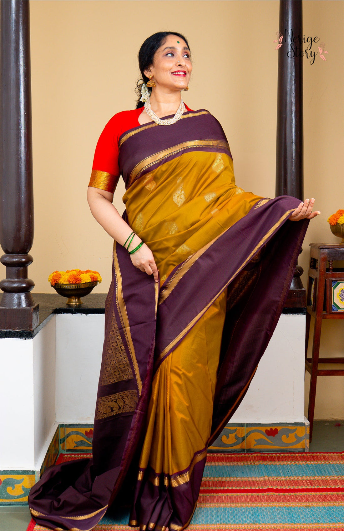 A woman modeling a traditional Indian saree in a studio setting. The saree has a deep purple border and pallu with a mustard yellow body, decorated with small gold motifs, and is paired with a bright red sleeveless blouse.